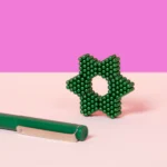 Ein stilisiertes, grünes, geometrisches Kunstobjekt aus kleinen Kugeln, das eine kreative und moderne Ästhetik vermittelt, vor einem rosa und lila Hintergrund, mit einem grünen Stift im Vordergrund.