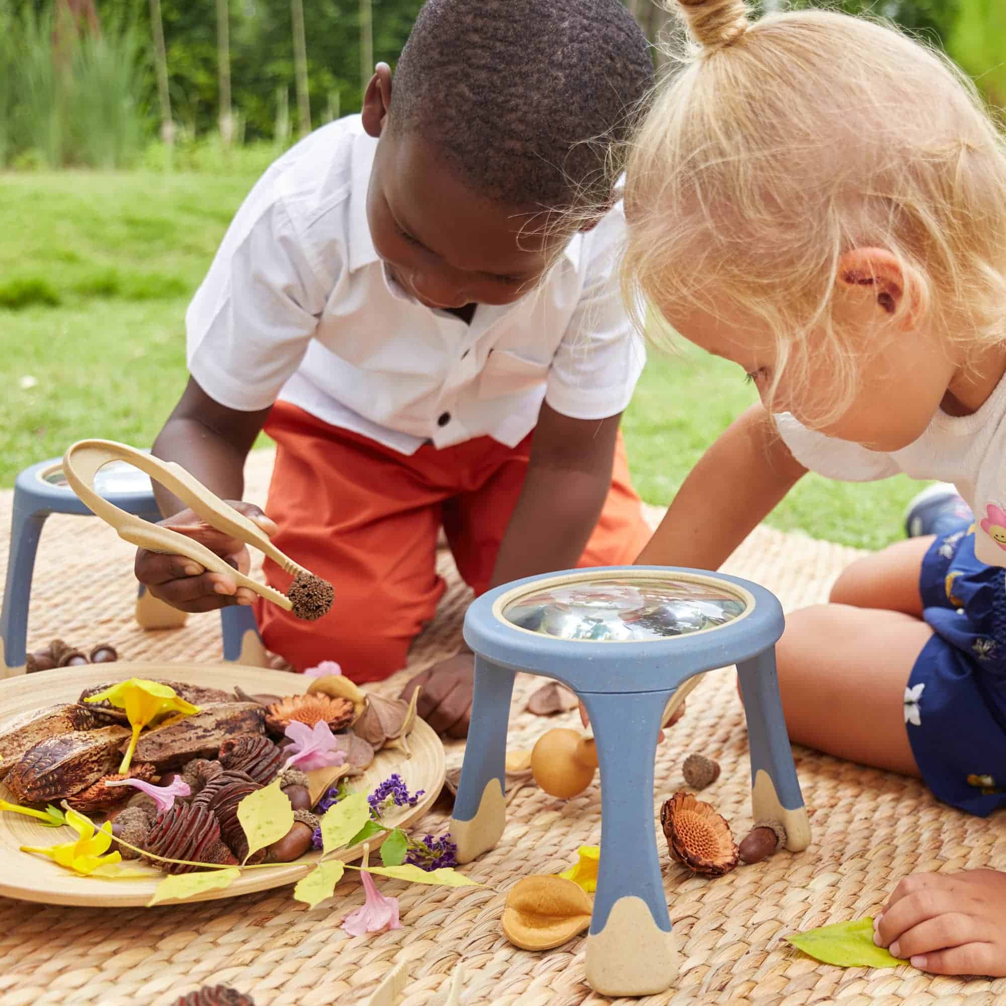 Im Bild sehen zwei Kinder, die draußen auf einer Decke mit Büchern und Naturmaterialien verschiedene Experimente durchführen und die Natur neugierig erkunden.