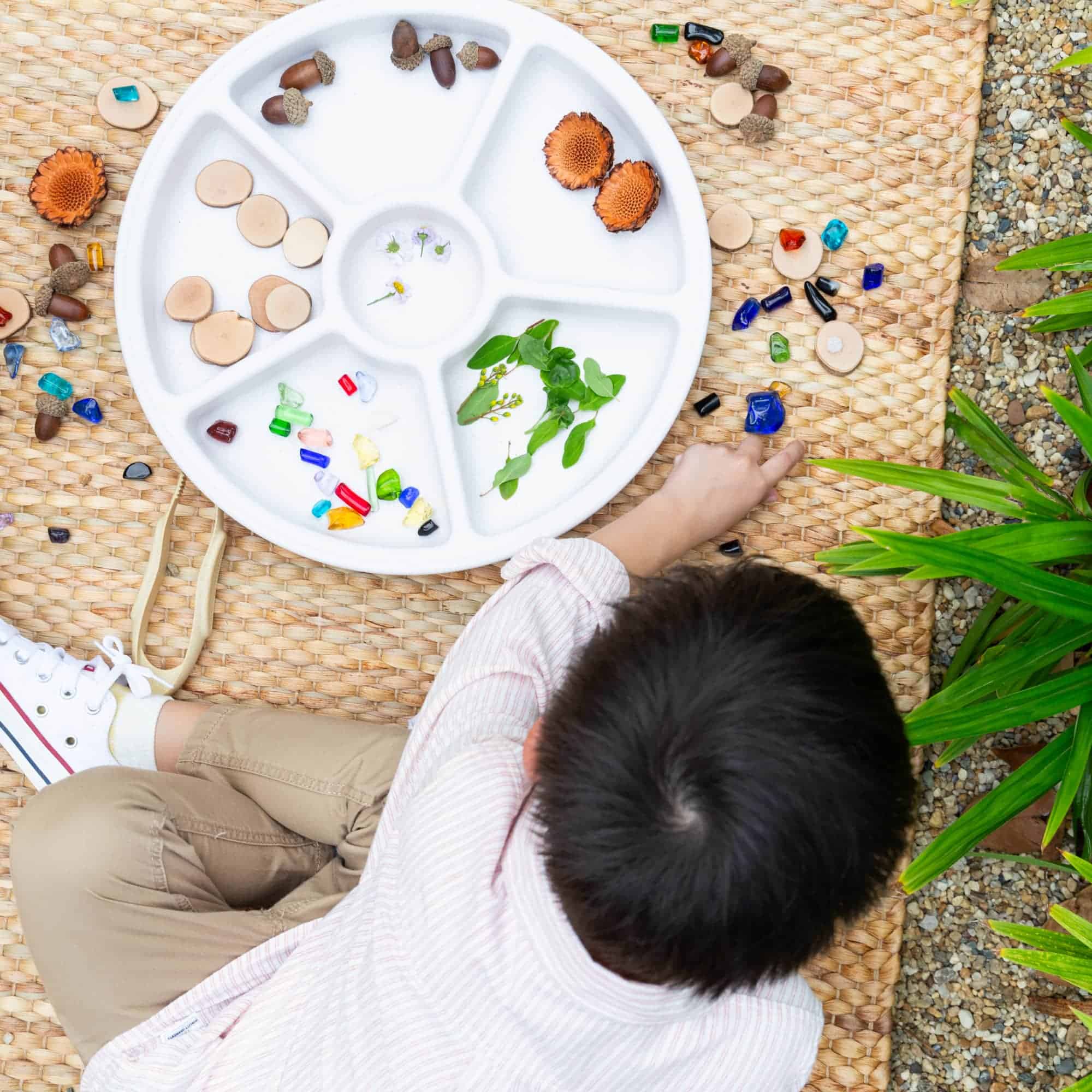 Verschiedene bunte Esskastanien, Blumen und Pflanzen auf einem fächerförmigen Maler- oder Basteltablett, das auf einem geflochtenen Untergrund außerhalb liegt. Ein Kind spielt mit den Materialien.