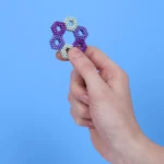 Bunter Perlenbrosche in Herzform, gehalten von einer Hand vor einem hellblauen Hintergrund.