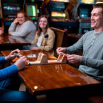 Entspannter Abend in einer Bar beim Spielen eines Brettspiels mit Freunden, während im Hintergrund Retro-Videospielautomaten leuchten.